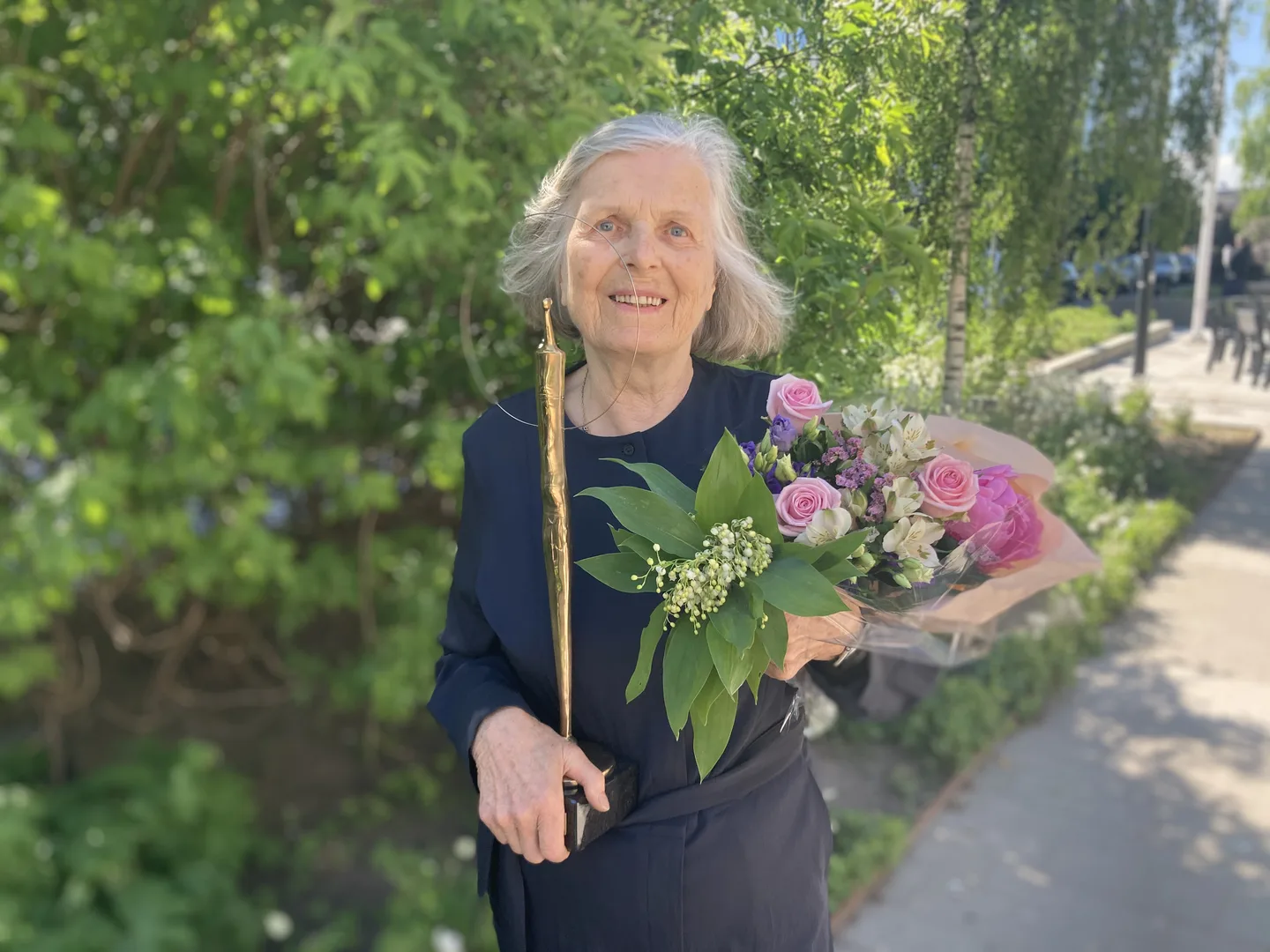 Karin Walde står i grönska med blombukett och håller Täby kommuns kulturpris i handen.