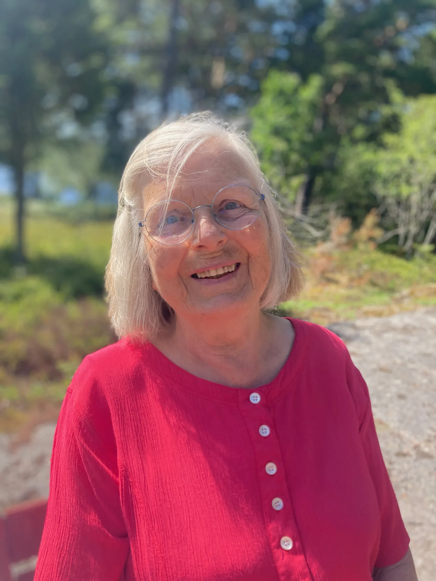 Närporträtt av Karin Walde i röd blus, leende i mjukt sommarljus med grön bakgrund.
