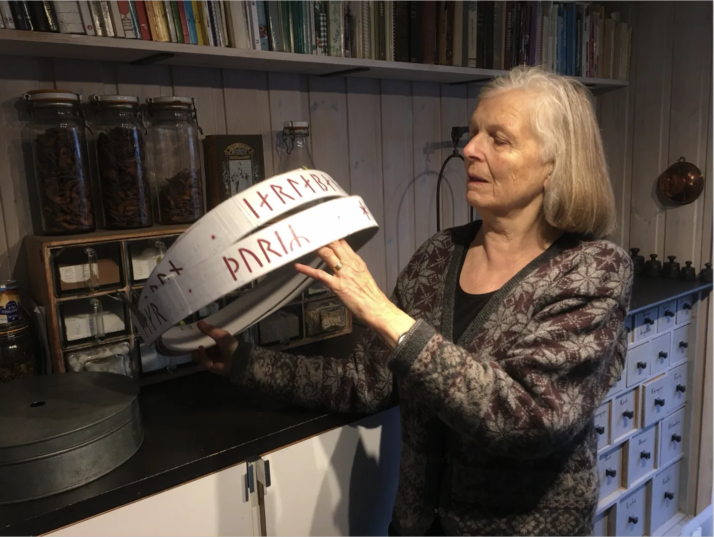 Karin Walde håller en vit modellring med röda runtecken framför hyllor med burkar och böcker.